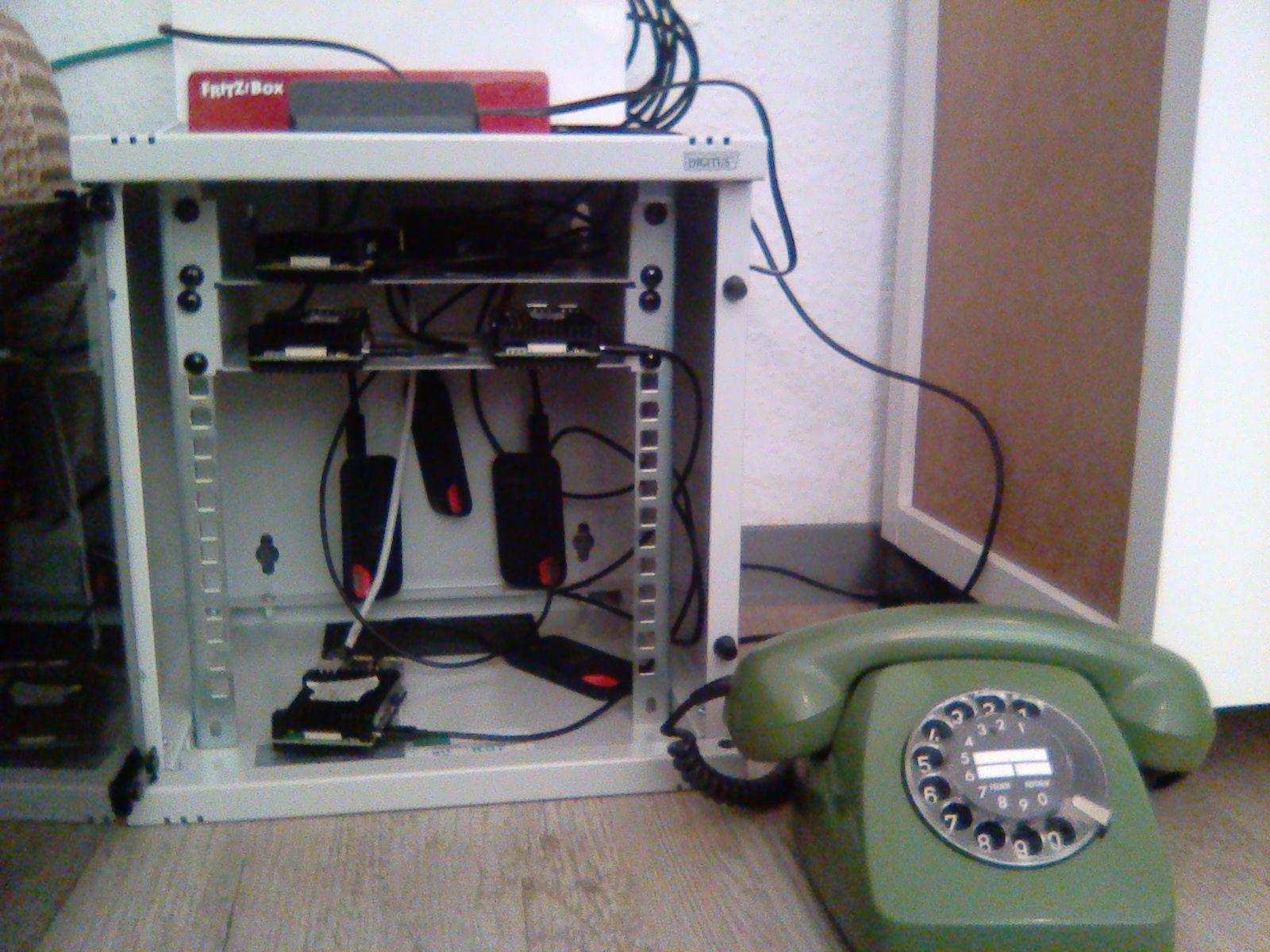 Ein kleines Netzwerk-Rack mit 4 Raspberry Pis darin, ein WLAN Router darauf und ein Telefon daneben